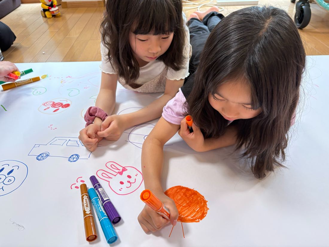 床に広げた紙にペンで絵を描く子どもたち